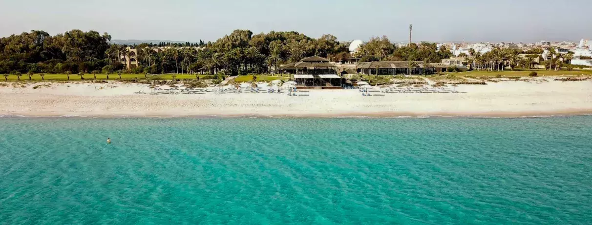 Plage de sable avec eau turquoise, transats et palmiers devant un resort entouré de verdure. 