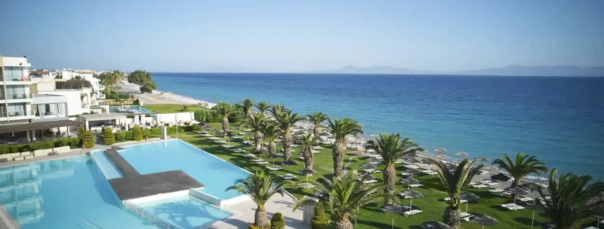 vue d'ensemble plage de galets, transats et parasols paillote, jardin, végétation, palmiers, chambres avec piscines partagées, hôtel avec bâtiments blancs