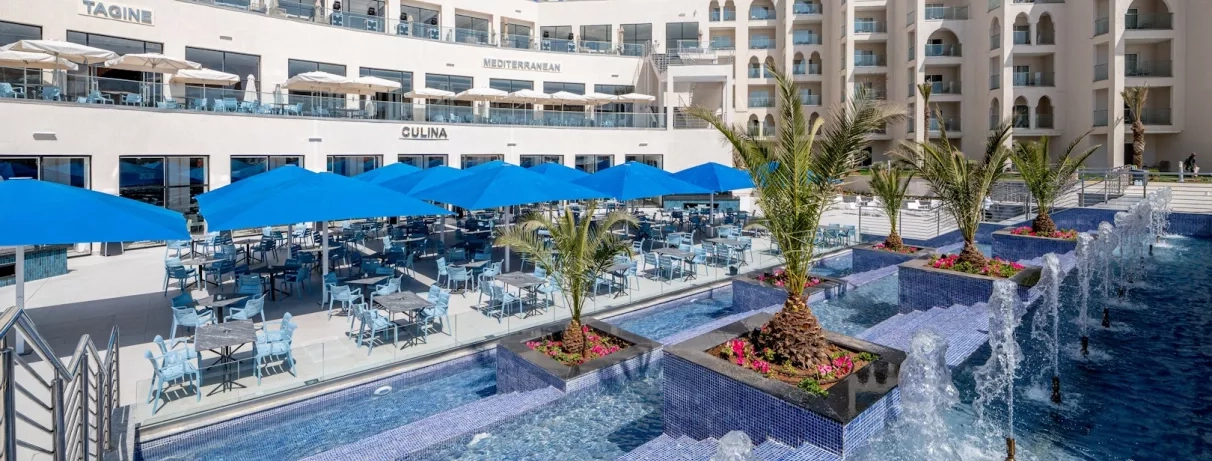 vue extérieure piscine palmiers fontaines jets à eau façade blanche hôtel paraols bleus