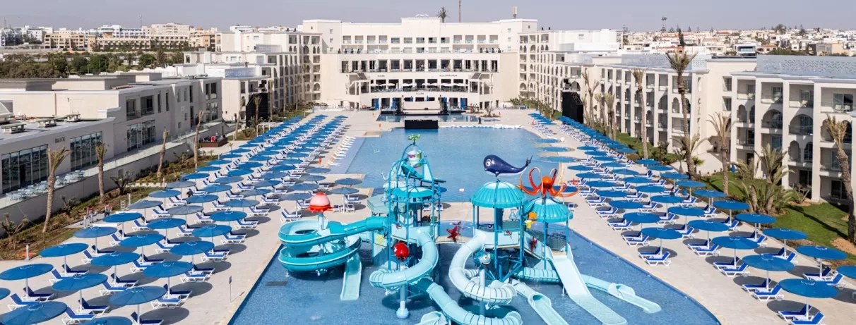 vue panoramique terrasse extérieure piscines tobogans bleus paraols et transats bleu blanc batiments blancs façade hôtel