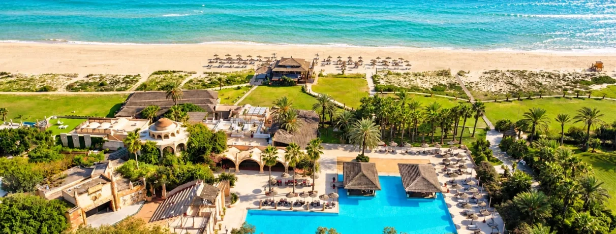 Vue aérienne d’un resort en bord de mer avec grande piscine, plage aménagée et palmiers.