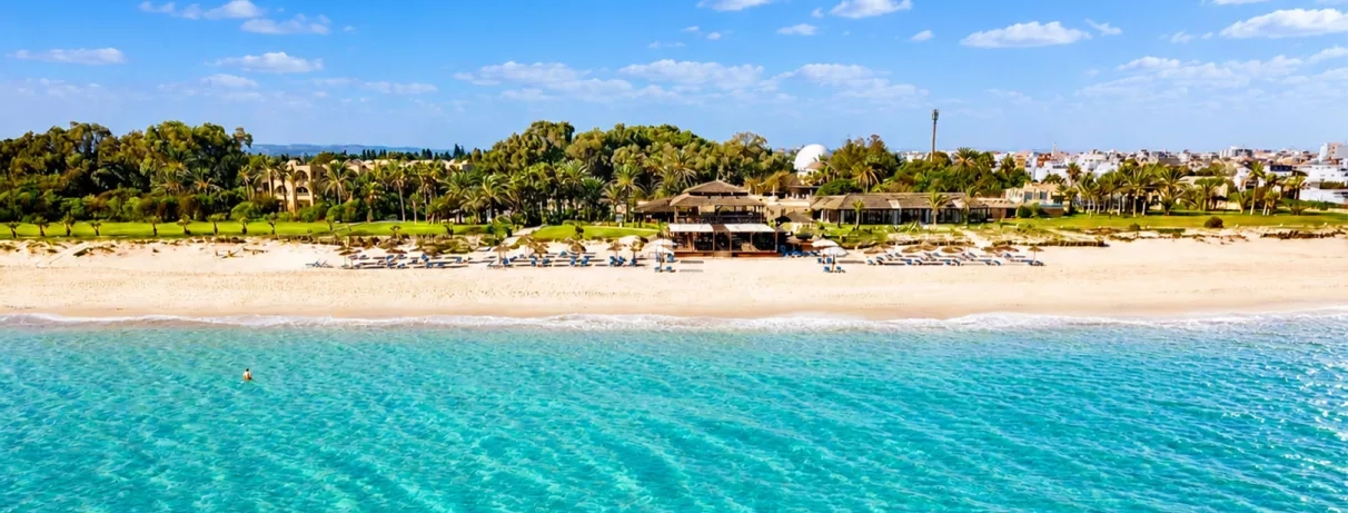 Plage de sable avec eau turquoise, transats et palmiers devant un resort entouré de verdure. 