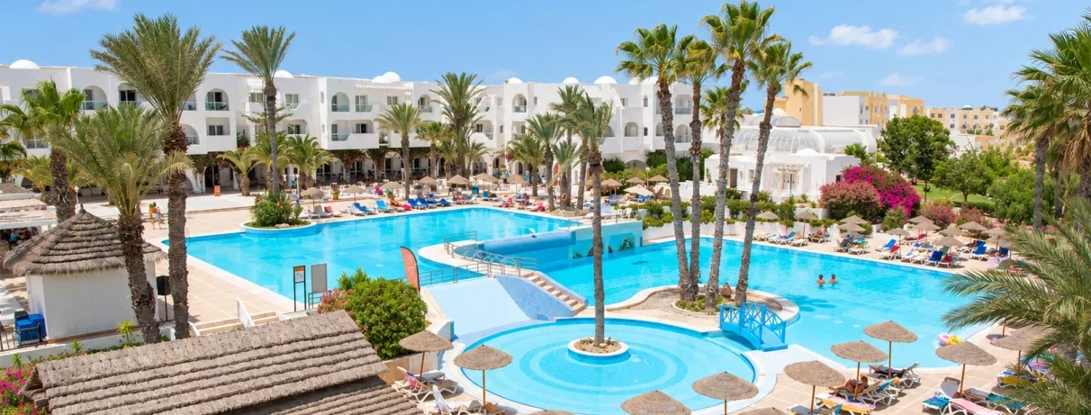 Vue panoramique de la grande piscine extérieure et des bâtiments blancs du Mondi Club Aladin à Djerba, bordée de palmiers et de transats.