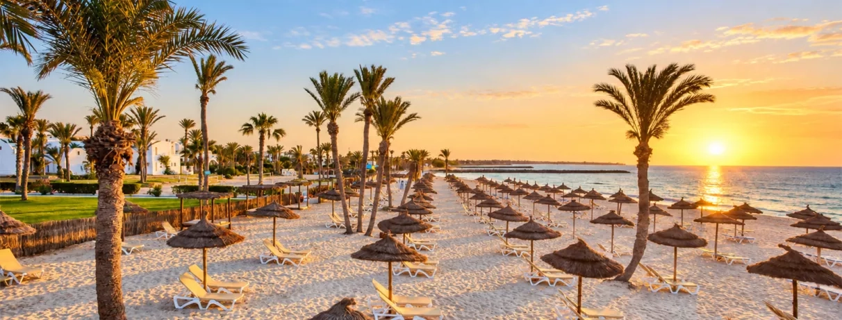 Plage privée du Mondi Club Aladin Djerba au coucher du soleil, alignement de parasols en paillote et transats face à la mer.