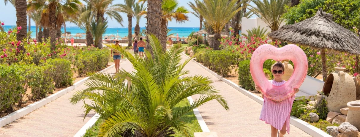 Petite fille tenant une bouée rose en forme de cœur sur une allée fleurie bordée de palmiers au Mondi Club Aladin Djerba.
