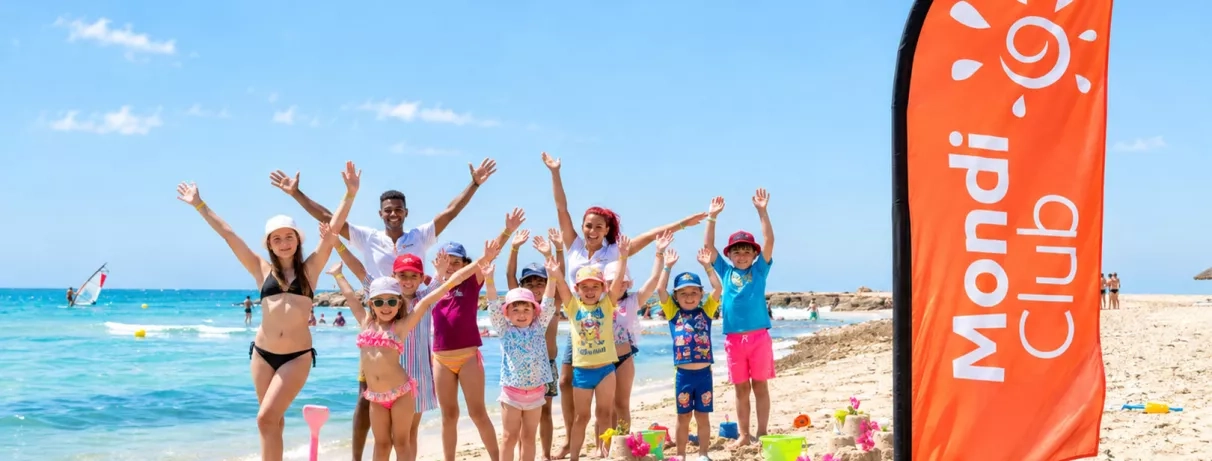 Enfants et animateurs du mini-club Mondi Club Aladin Djerba levant les bras sur la plage devant l'oriflamme du club.