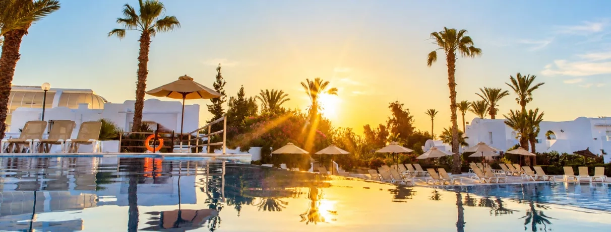 Coucher de soleil se reflétant dans la piscine extérieure entourée de palmiers et de transats au Mondi Club Aladin Djerba.