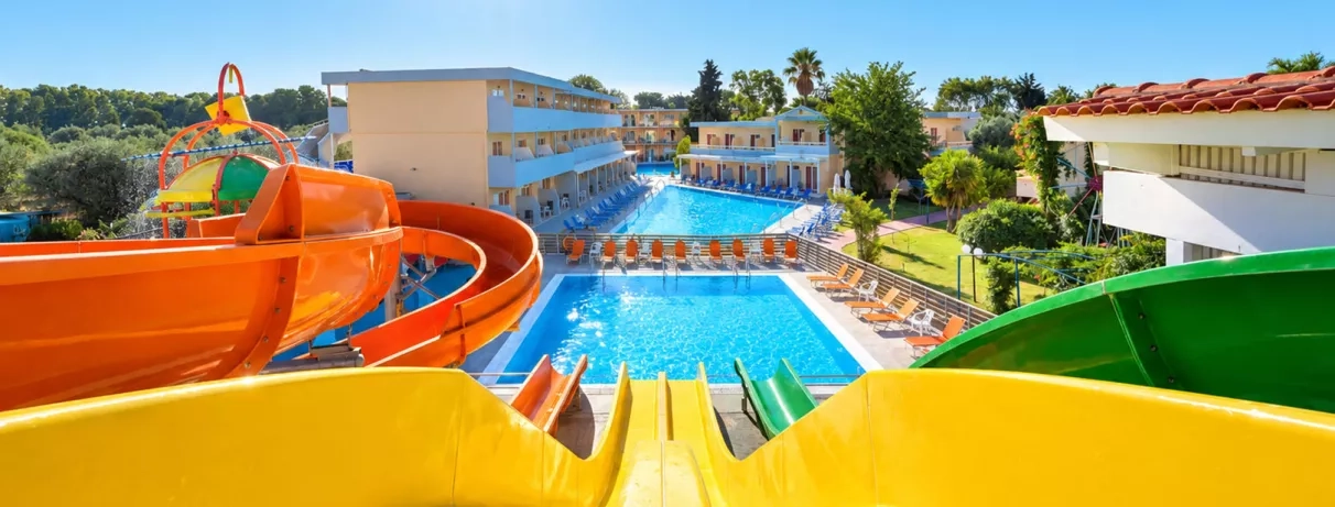 toboggans, deux piscines et transats autour, végétation, plusieurs bâtiments avec deux étages, ciel bleu