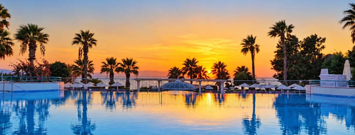 Un coucher de soleil flamboyant se reflétant sur l'eau de la piscine bordée de palmiers.