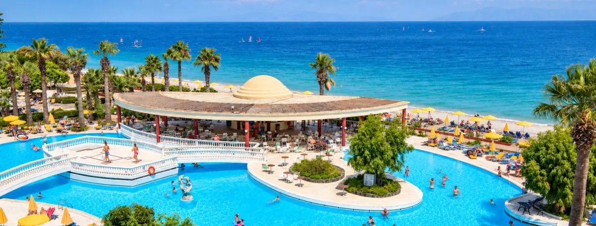 vue aérienne mer et piscine, bar de la piscine, pont et plateforme permettant de traverser la piscine, accès plage de galets, palmiers, transats et parasols