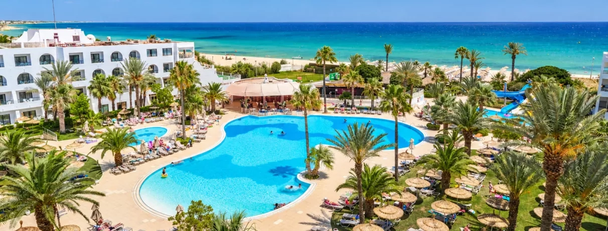 Vue panoramique sur la piscine lagune du Mondi Club Nozha Beach à Hammamet avec palmiers, transats et vue sur la mer Méditerranée.