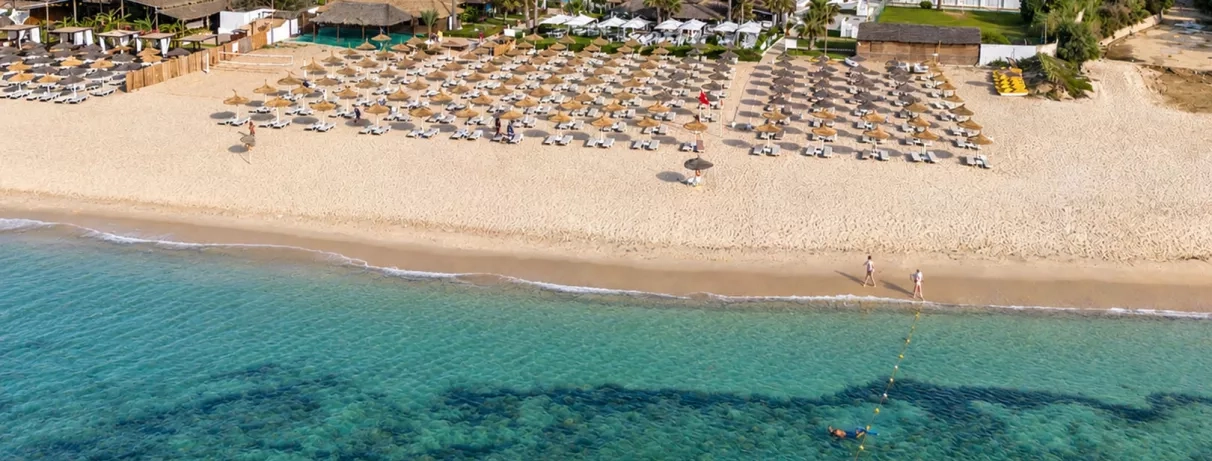 Vue aérienne de la plage privée aménagée avec parasols en paille et mer cristalline au Mondi Club Nozha Beach Hammamet.