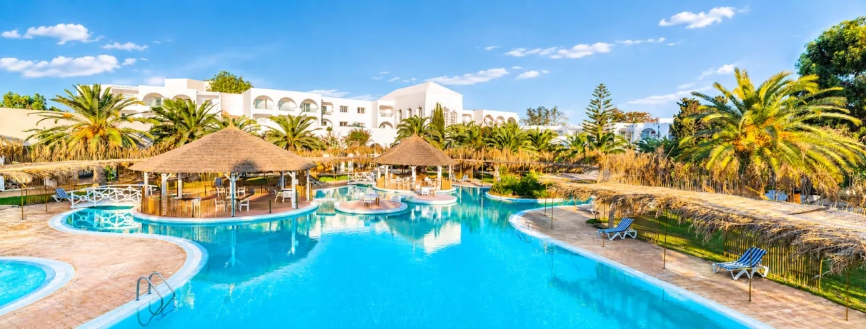 Grande piscine lagon du Mondi Club Shalimar Hammamet bordée de palmiers et d'aillottes en paille.