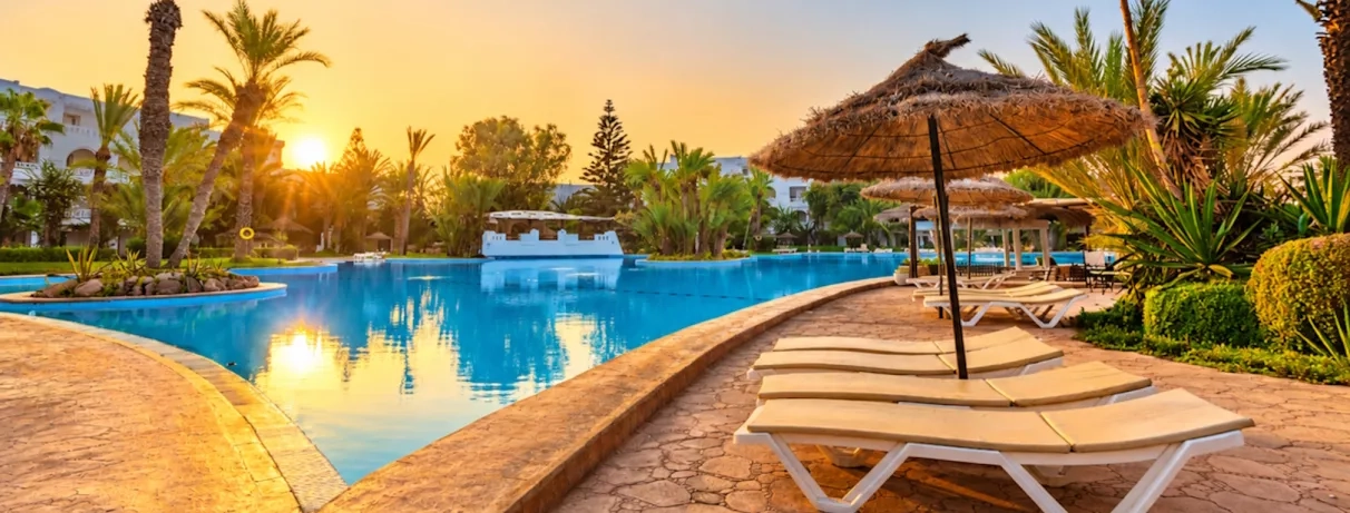 Grande piscine extérieure au coucher du soleil avec transats et parasols de paille au Djerba Resort.