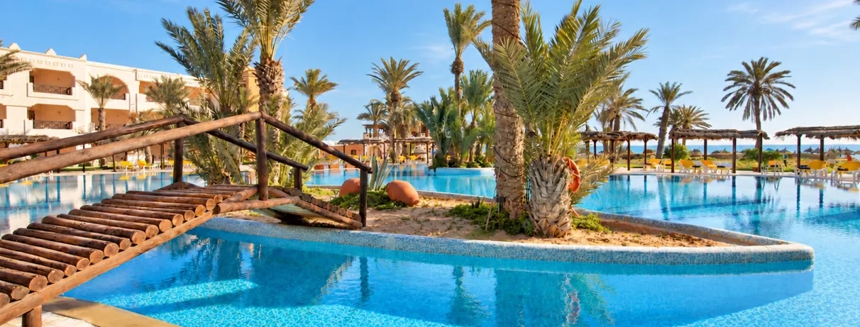 Grande piscine lagon bordée de palmiers et pont en bois sous le soleil de l'hôtel Safira Palms Djerba.
