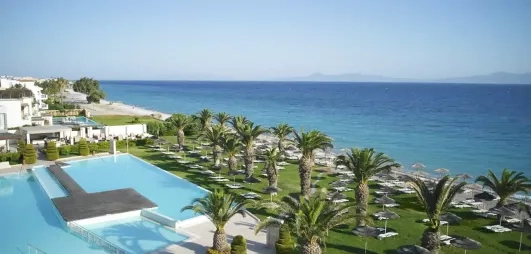 vue d'ensemble plage de galets, transats et parasols paillote, jardin, végétation, palmiers, chambres avec piscines partagées, hôtel avec bâtiments blancs