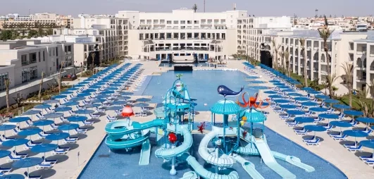 vue panoramique terrasse extérieure piscines tobogans bleus paraols et transats bleu blanc batiments blancs façade hôtel