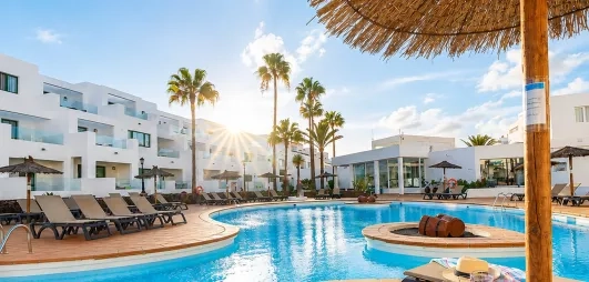 Une vue panoramique d'une grande piscine, bordée de transats et de bâtiments blancs modernes.