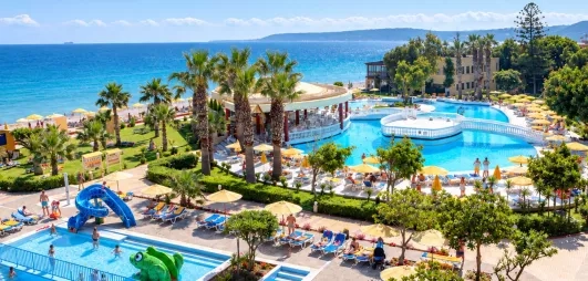 vue aérienne piscines, petit toboggan dans la piscine enfant, palmiers, pont et plateforme sur la piscine adulte, accès plage de galets, pieds dans l'eau, transats 