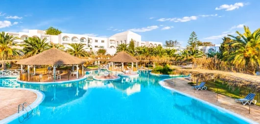 Grande piscine lagon du Mondi Club Shalimar Hammamet bordée de palmiers et d'aillottes en paille.