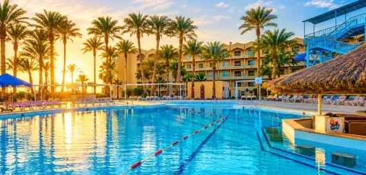 vue piscine extérieur avec bar central, transats autour, coucher de soleil, palmier, bâtiment de l'hôtel