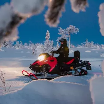 Une motoneige rouge avec phares allumés circulant sur une piste de neige entre des sapins givrés à Sodankylä.