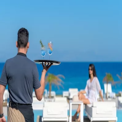 Serveur avec deux cocktails frais, au bord de la piscine, vue sur plage, transats 