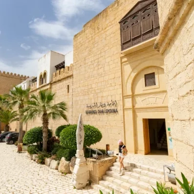 Un bâtiment en pierre de style méditerranéen, avec des palmiers et une entrée marquée “Résidence Diar Lemdina”, devant lequel une personne descend quelques marches sous un ciel ensoleillé.