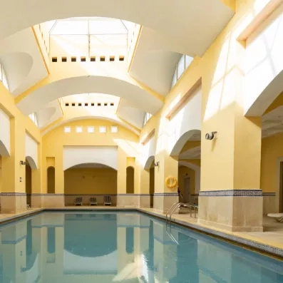 Une piscine intérieure lumineuse, entourée d’arches élégantes et de murs jaune clair, est baignée de lumière naturelle provenant d’un plafond vitré.
