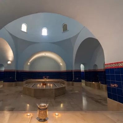 Une salle de hammam élégante à l’architecture voûtée, éclairée par de petites ouvertures en hauteur, avec une fontaine octogonale au centre entourée de bougies sur un sol en marbre.