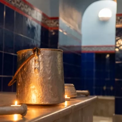 Un seau métallique éclairé par une lueur chaleureuse de bougies repose sur un banc en marbre dans un hammam aux murs carrelés de bleu profond et aux arches élégantes.