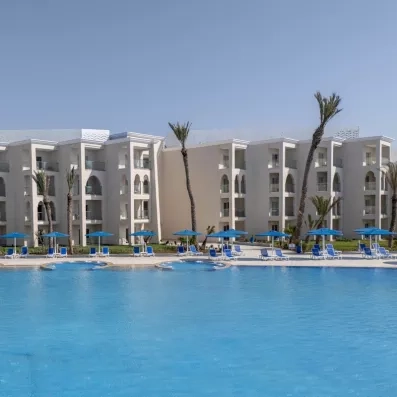 vue de loin façade hôtel blanc piscine transats et parasols bleus long palmiers