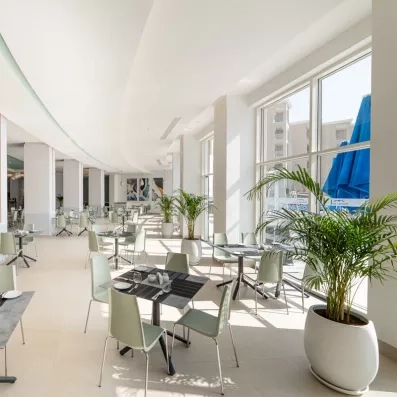 grande salle blanche tables et chaises blanches grises plantes murs blancs et parasols bleus dehors par la fenêtre