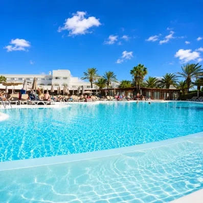 Une grande piscine d'hôtel s'étend sous un ciel bleu, bordée de palmiers.