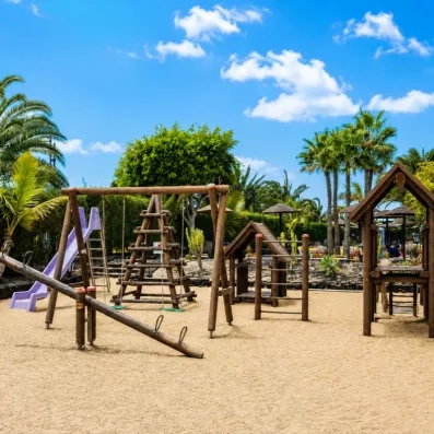 Une aire de jeux pour enfants en bois installée sur un sol souple, entourée de palmiers et de verdure.
