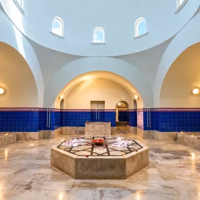 Une salle de hammam élégante à l’architecture voûtée, éclairée par de petites ouvertures en hauteur, avec une fontaine octogonale au centre entourée de bougies sur un sol en marbre.