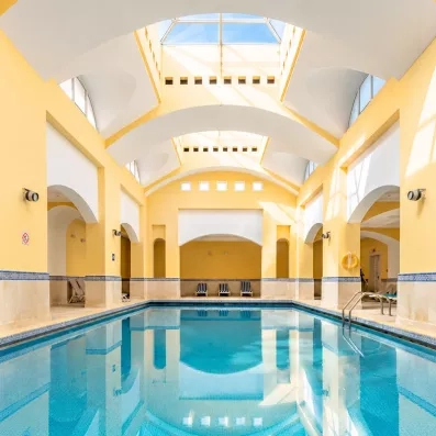 Une piscine intérieure lumineuse, entourée d’arches élégantes et de murs jaune clair, est baignée de lumière naturelle provenant d’un plafond vitré.