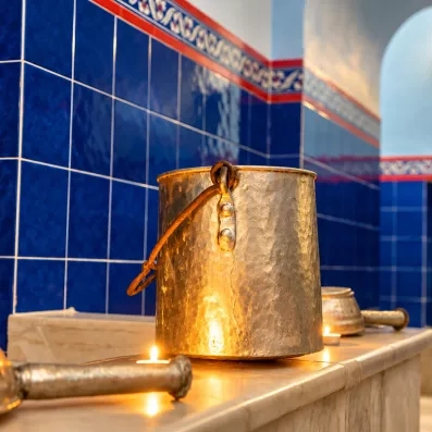 Un seau métallique éclairé par une lueur chaleureuse de bougies repose sur un banc en marbre dans un hammam aux murs carrelés de bleu profond et aux arches élégantes.