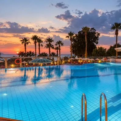 Une grande piscine extérieure, entourée de palmiers sous un ciel aux teintes rosées.