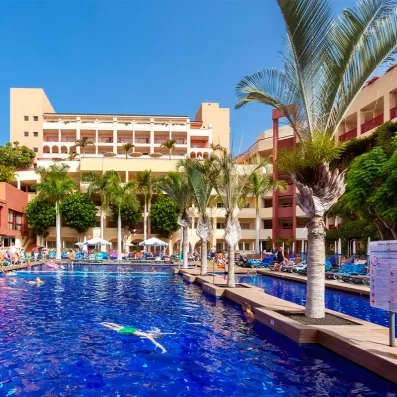 Une piscine extérieure ensoleillée, entourée de palmiers et de vacanciers.