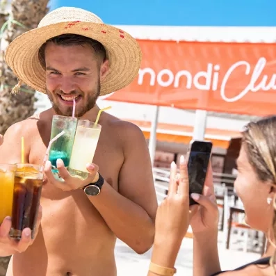 homme avec chapeau et cocktails et une femme qui le prend en photo avec son téléphone, bannière mondi club en arrière plan