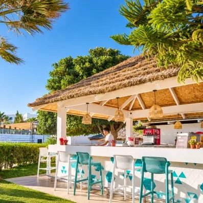 Bar extérieur en plein air abrité sous un toit de chaume avec tabourets hauts turquoise, situé dans les jardins tropicaux du complexe hôtelier.