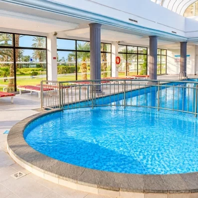Piscine intérieure couverte du Mondi Club One Resort Jockey à Monastir, entourée de colonnes et de baies vitrées donnant sur le jardin.