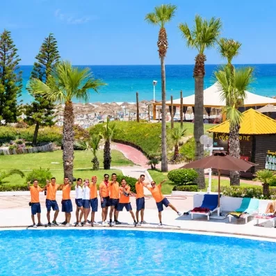 Équipe d'animation du Mondi Club One Resort Jockey posant au bord de la piscine avec palmiers et mer Méditerranée en arrière-plan.
