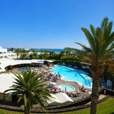 Une vue montre une grande piscine d'hôtel entourée de palmiers et de transats.