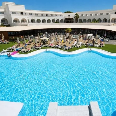 Une vue aérienne montre une grande piscine entourée de transats et d'un bâtiment d'hôtel blanc en arrière-plan.
