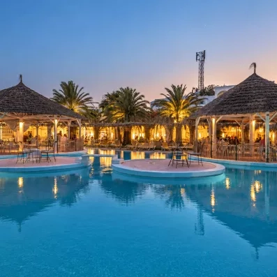 Grande piscine extérieure éclairée au crépuscule avec paillotes au Mondi Club Shalimar.
