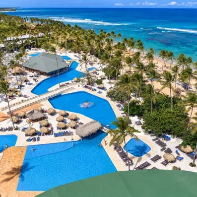 piscines avec accès direct sur la plage de sable, palmiers, végétation luxuriante, transats bleus et parasols paillote, pieds dans l'eau, océan et ciel bleus