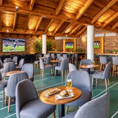 salle de restaurant chaises grises et tables rondes en bois, bière et burgers posés sur une table, toit en bois poutres apparentes, écrans diffusant du sport au mur
