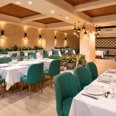 salle de restaurant chaises vertes longues tables nappes blanches, pilier et luminaires au centre, beaucoup de plantes 